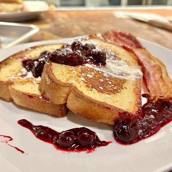 French toast with berry compote, powdered sugar, and a side of bacon on a white plate.